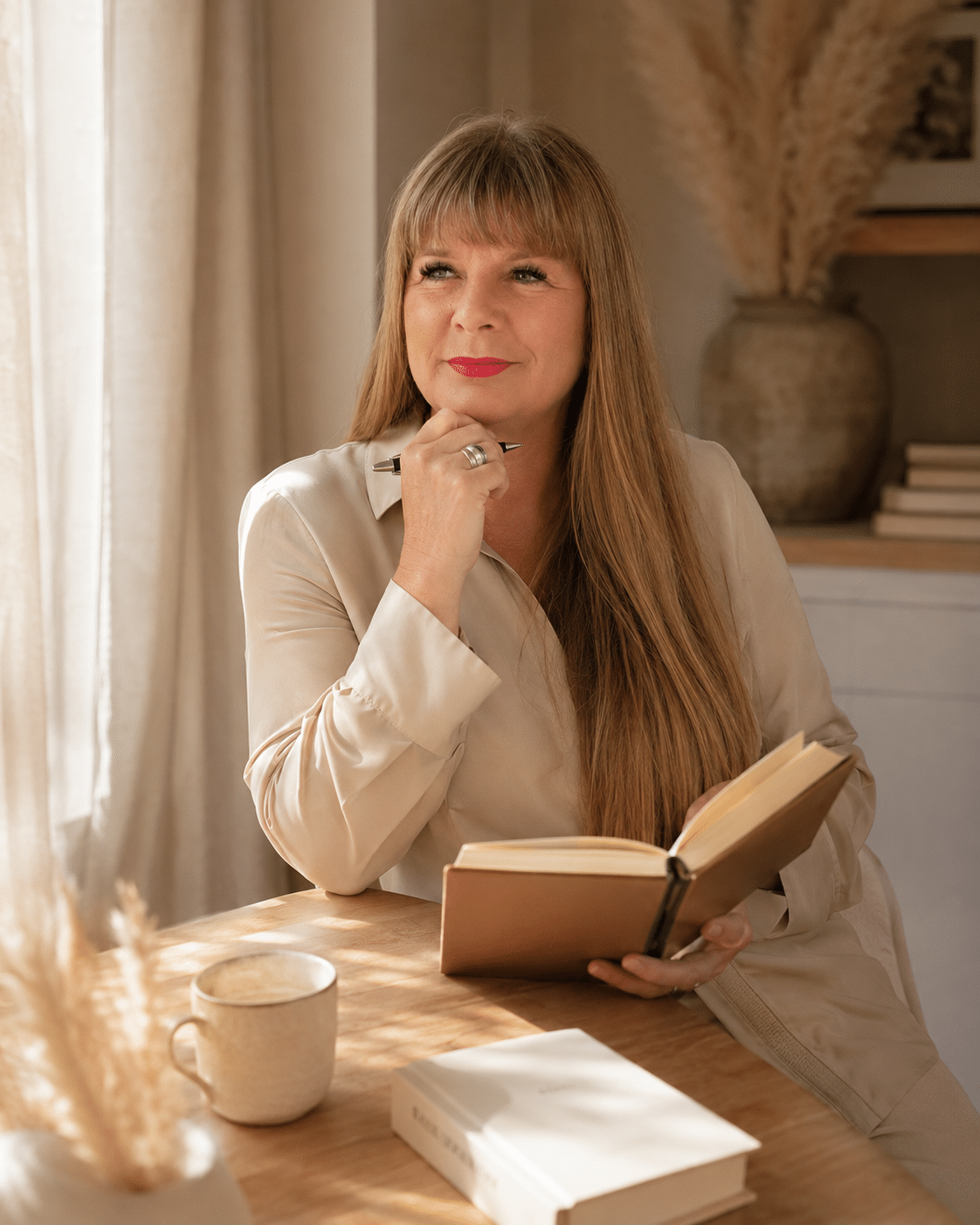 Anette Pelzer sitzt in warmer, heller Atmosphäre mit einem geöffneten Buch am Tisch und blickt nachdenklich in die Zukunft von Menschenkompass.
